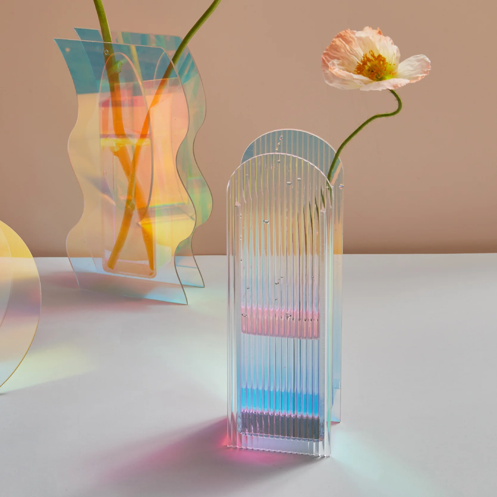 vase acrylique iridescent à motifs nervurés, posé sur une table blanche. Le vase ultra épais reflète des couleurs arc-en-ciel et projette un jeu de lumière coloré, parfait pour une décoration bohème chic.