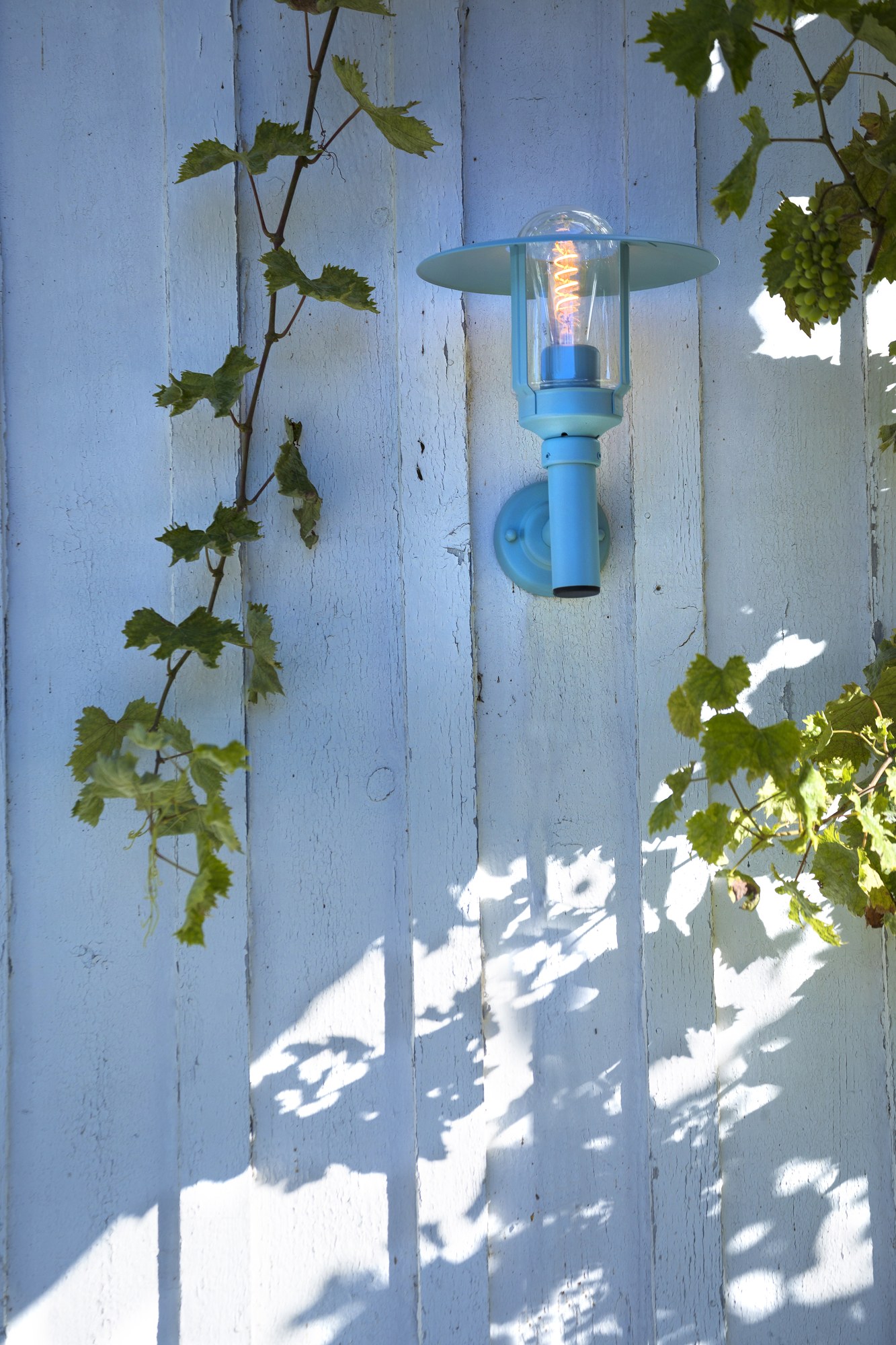 Applique montante Newpark par Roger Pradier en aluminium applique montante Newpark en aluminium bleu fixée sur un mur de planches peintes sous une vigne