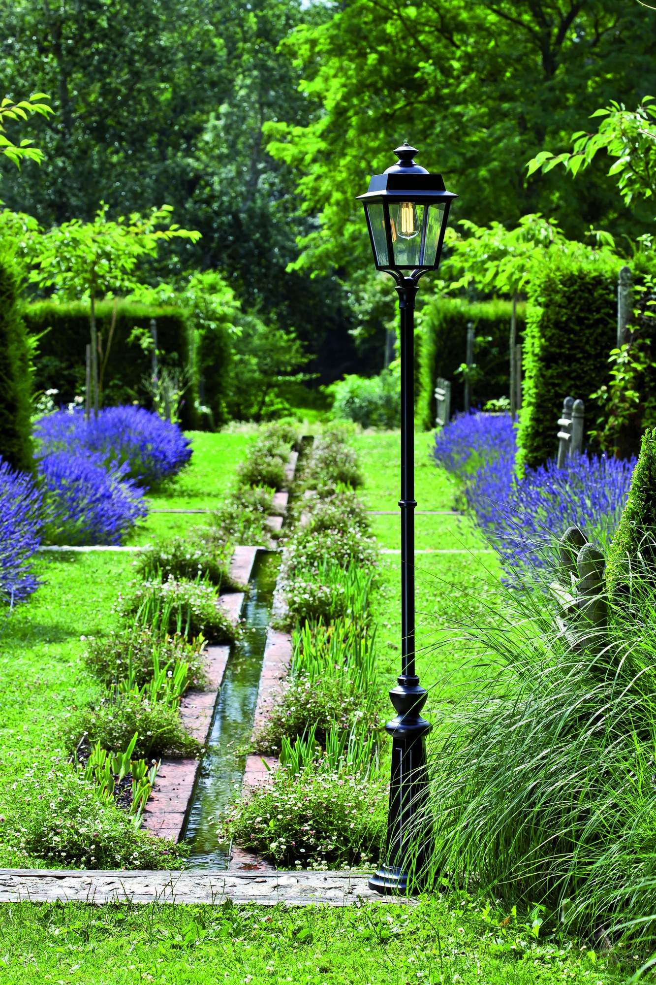 Lampadaire Place des Vosges Évolution en aluminium par Roger Pradier lampadaire Place des Vosges Évolution colonne ornée haute dans une allée de jardin bordée de lavande