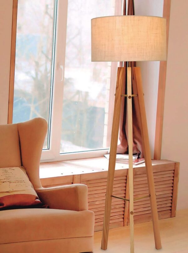 lampadaire trépied en bois allumé dans un salon, devant une fenêtre avec un fauteuil à côté