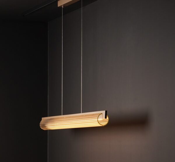 Suspension Lirra 130 LED allumée devant un mur sombre, finition laiton