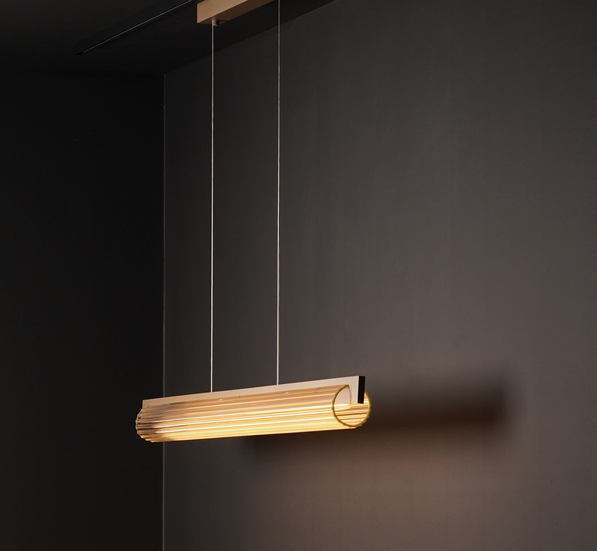 Suspension Lirra 130 LED allumée devant un mur sombre, finition laiton