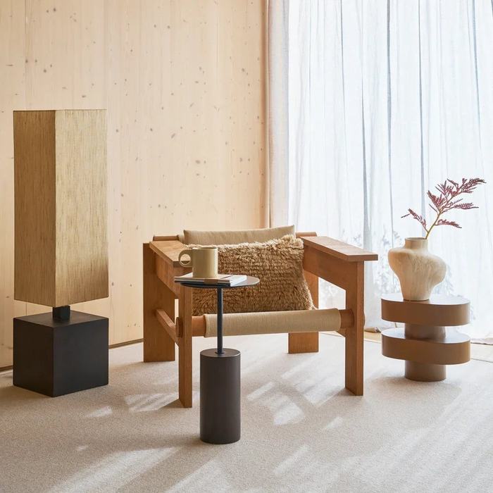 Table d'appoint bois : laquelle choisir pour votre salon ? Table d'appoint bois Macy A à socle en manguier et plateau en métal noir devant un fauteuil en bois