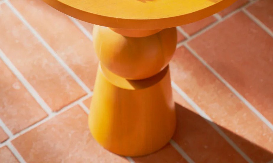 Table d'appoint Teko par Urban Nature Culture, manguier orange doré table d'appoint bois Teko Urban Nature Culture, sur un sol en briques terracotta avec des livres sur le plateau
